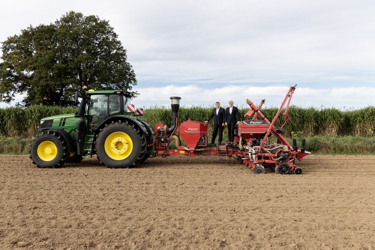 Two men are standing on a seeder attached to a tractor.